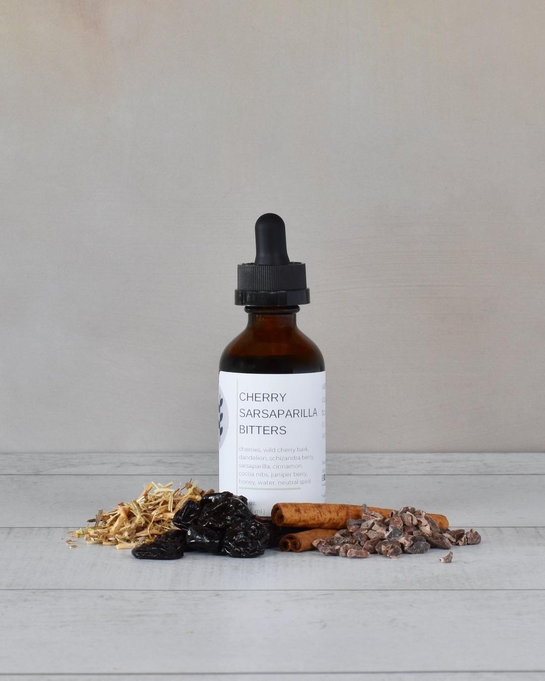 FreeWildShe Cherry Sarsaparilla Bitters sit on a white countertop surrounded by ingredients.