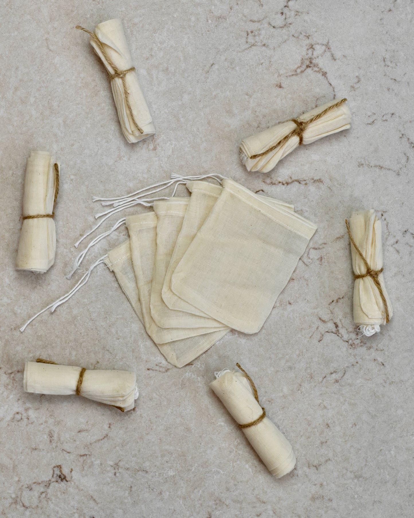 Five FreeWildShe Reusable Cotton Tea Bags lay spread out in the center on a cream colored countertop. Rolled bundles wrapped in twine lay scattered around.