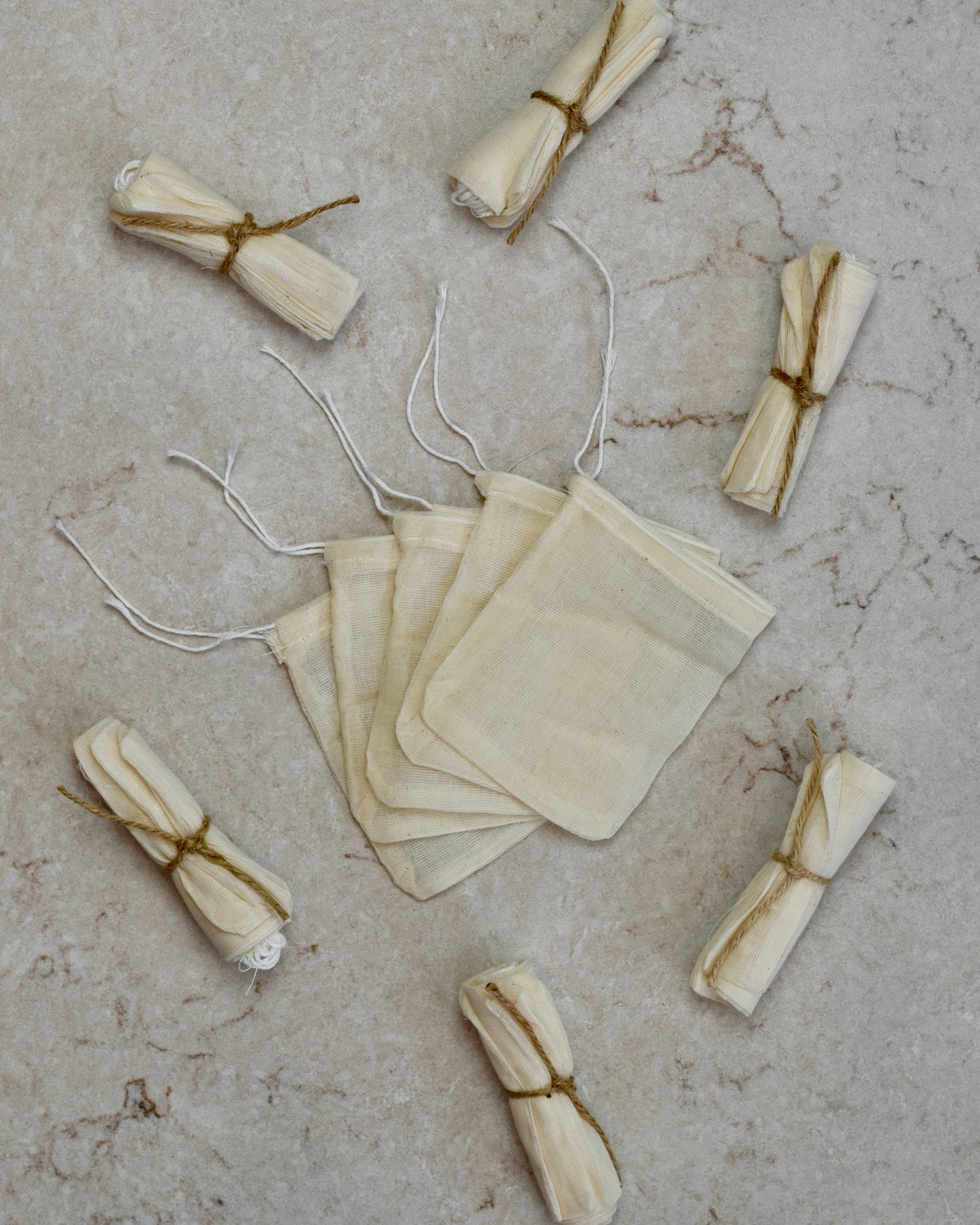 Five FreeWildShe Reusable Cotton Tea Bags lay spread out in the center on a cream colored countertop. Rolled bundles wrapped in twine lay scattered around.