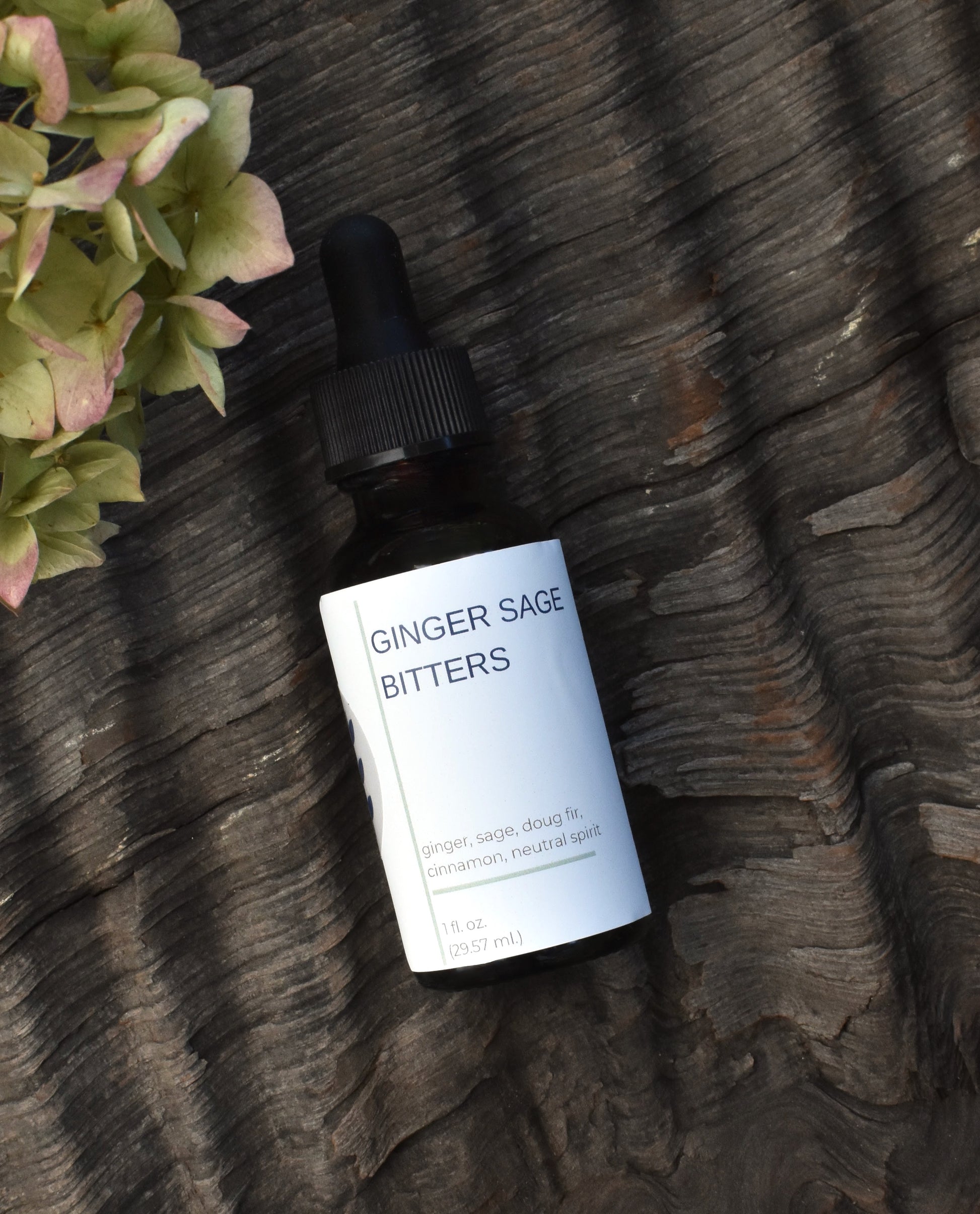 FreeWildShe Ginger Sage Bitters lay on a textured piece of bark surrounded by dried hydrangea blooms.