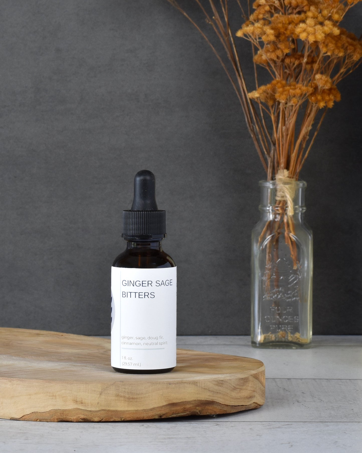FreeWildShe Ginger Sage Bitters sit on a wooden platter, on a white wooden table. A bouquet of dried flowers is in the background.
