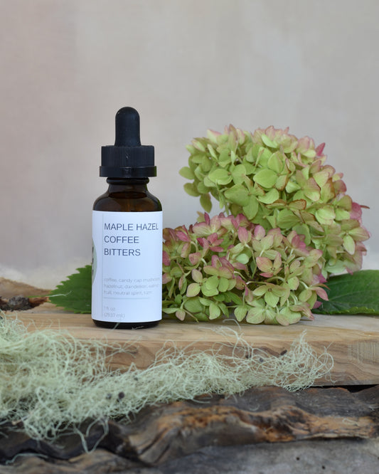 FreeWildShe Maple Hazel Coffee Bitters sit on a wooden tray. Lichen and hydrangea blooms surround the image.