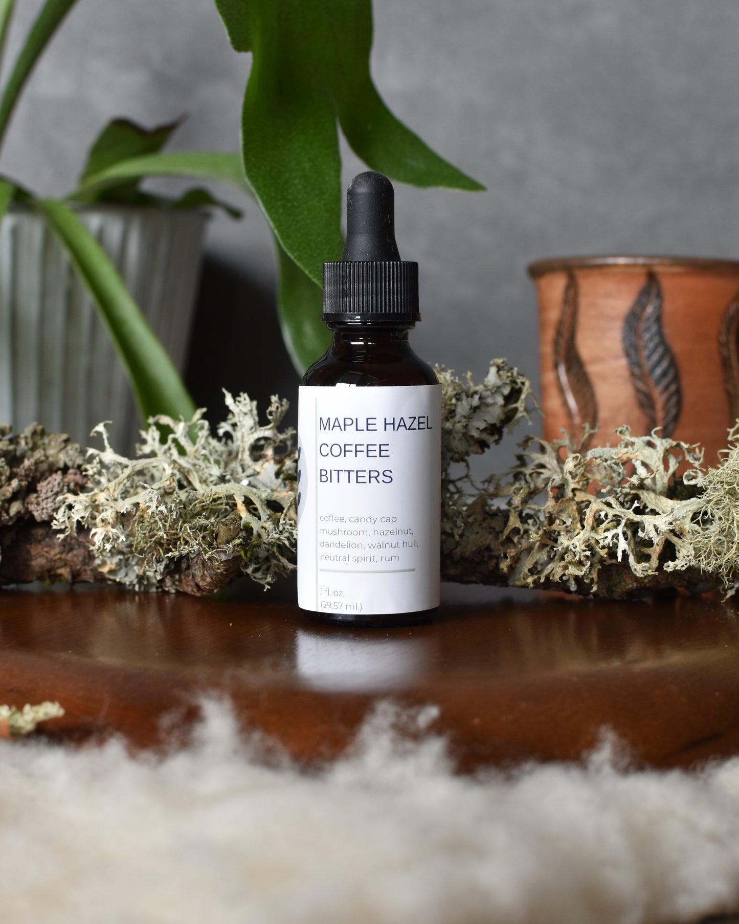 FreeWildShe Maple Hazel Coffee Bitters sit on a wooden plate, atop a fuzzy wool blanket. Lichen, a mug, and a houseplant sit in the background.