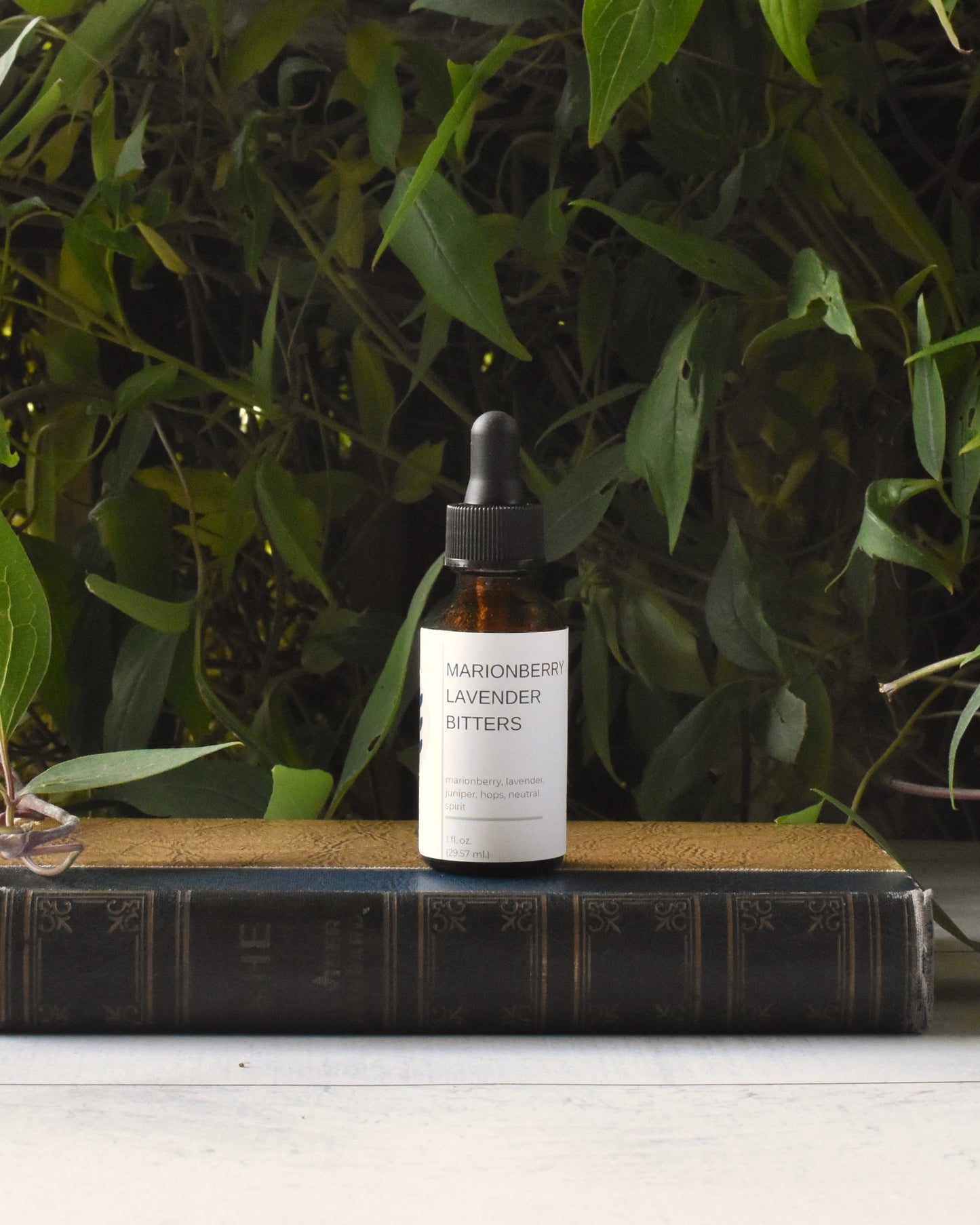 FreeWildShe Marionberry Lavender Bitters sit on an old book, in front of a green vine.
