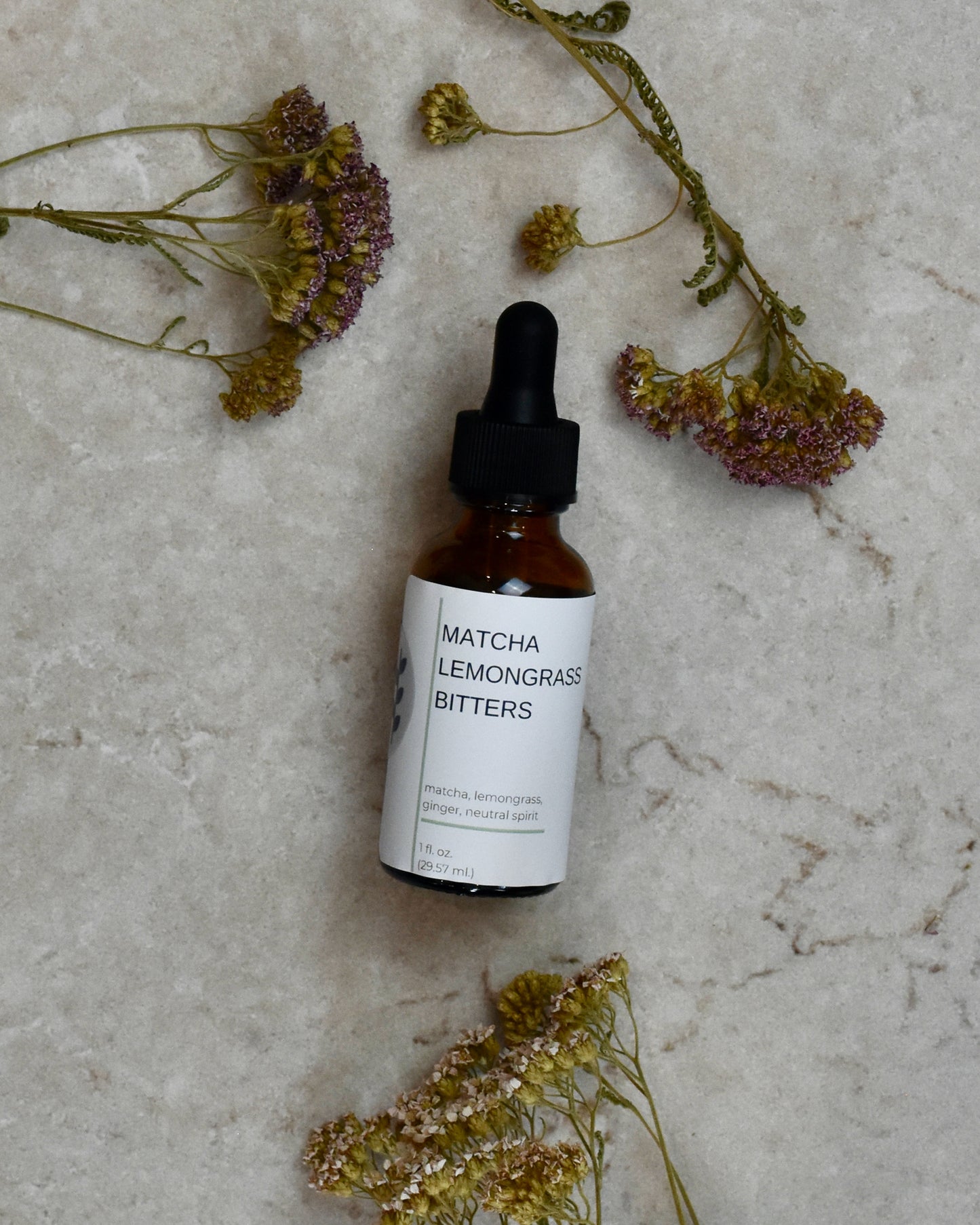 FreeWildShe Matcha Lemongrass Bitters lay on a cream colored countertop. Dried flowers are scattered around.