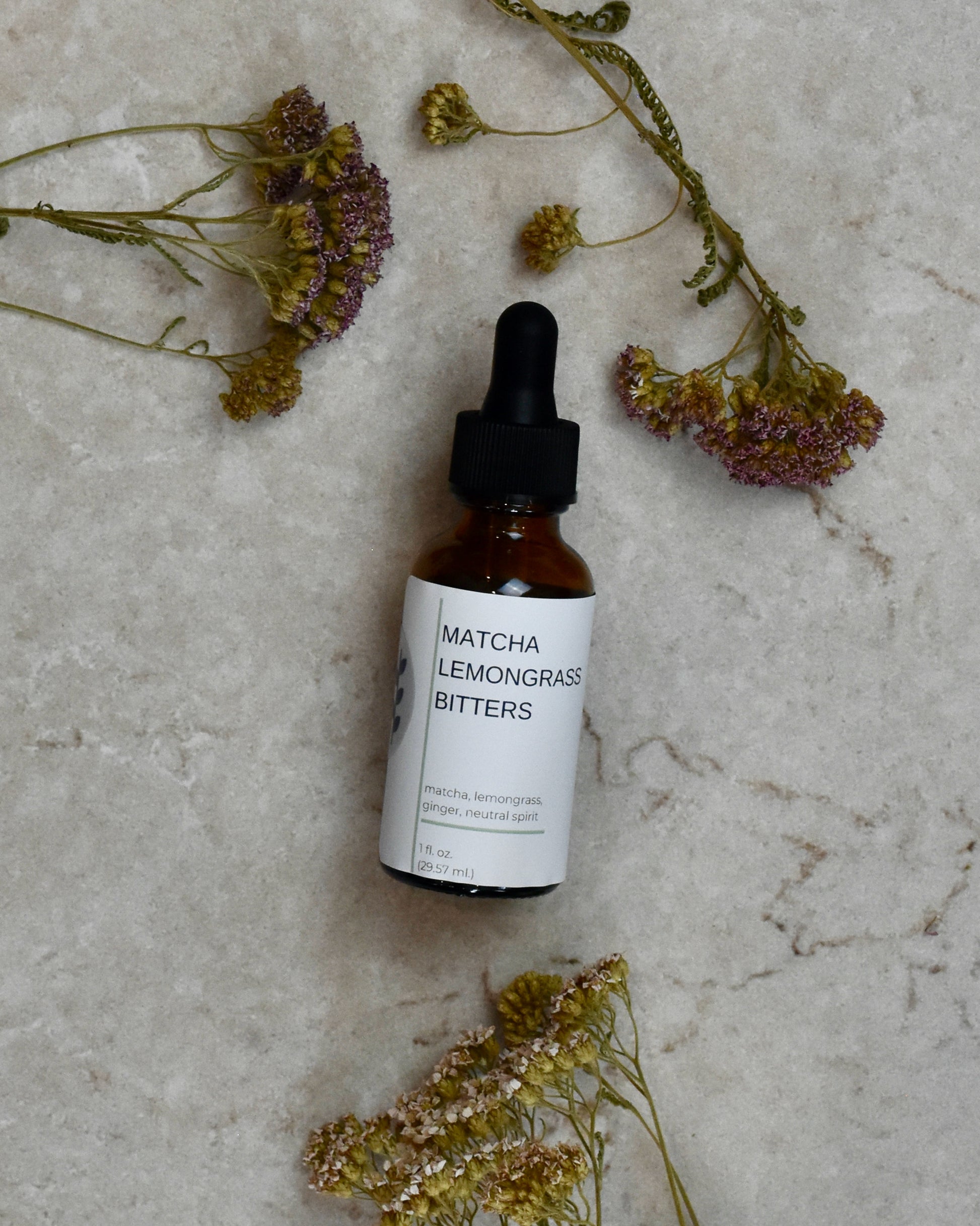 FreeWildShe Matcha Lemongrass Bitters lay on a cream colored countertop. Dried flowers are scattered around.