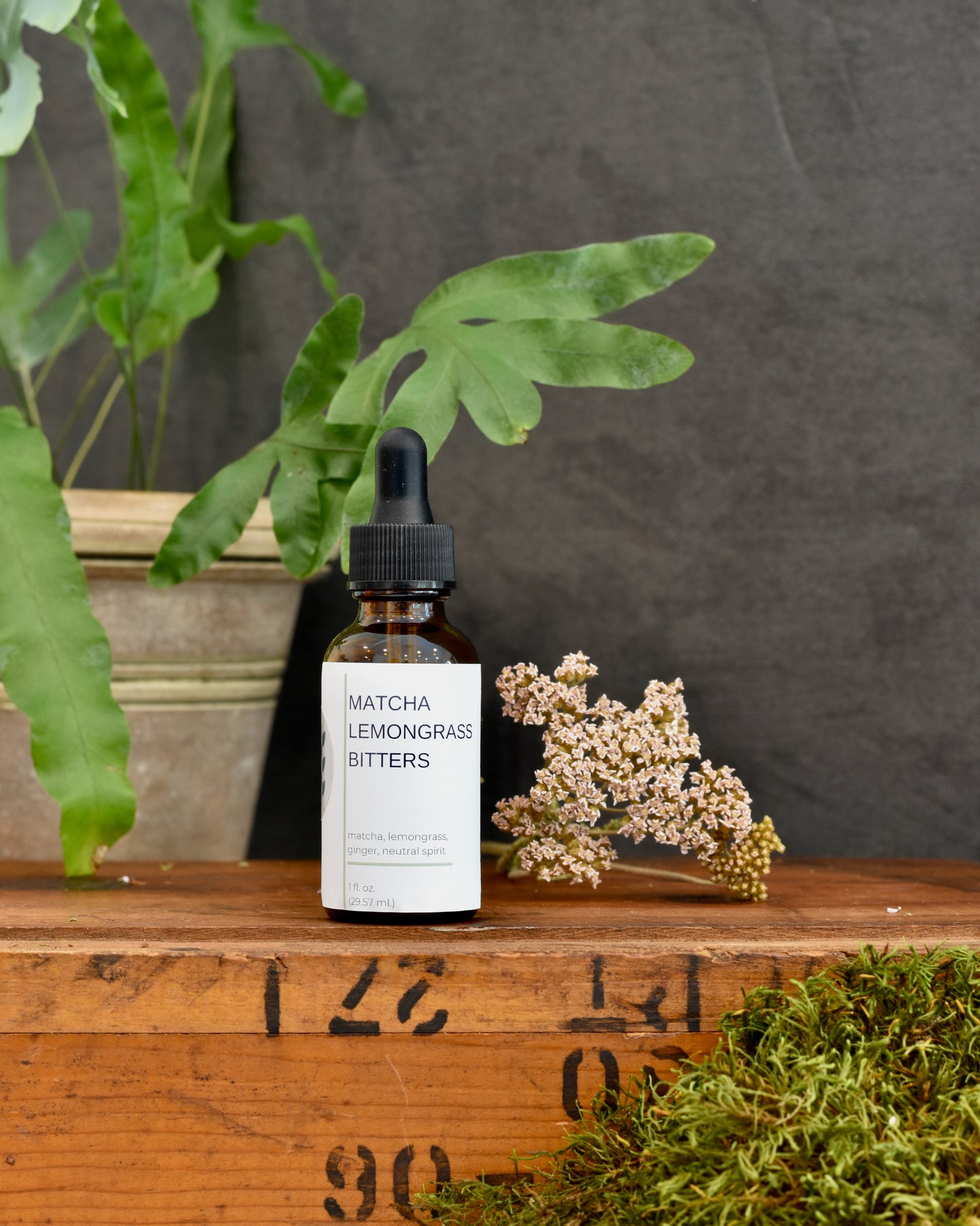 FreeWildShe Matcha Lemongrass Bitters sit on a wooden box. A plant and dried flower sit in the background.