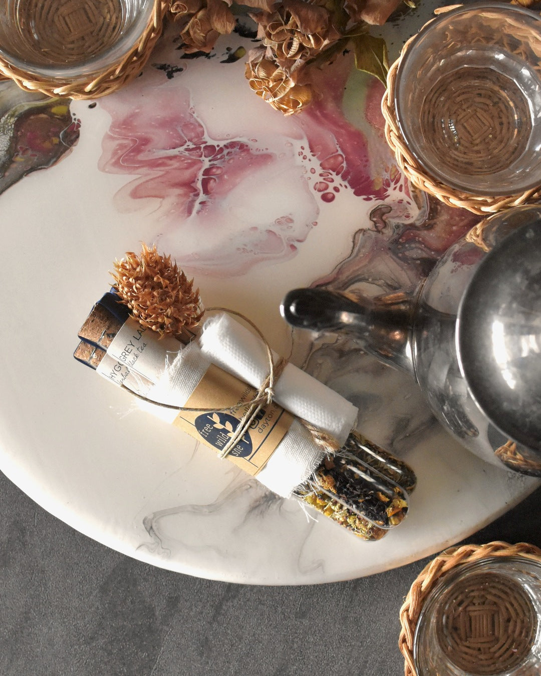 Three FreeWildShe tea vials are bundled together with a wrap of muslin, kraft paper, & twine. A sprig of dried clover is tucked into the bundle. The Mother's Mix tea gift pack sits on a platter surrounded by glass & wicker tea mugs, a metal teapot, and dried flowers.