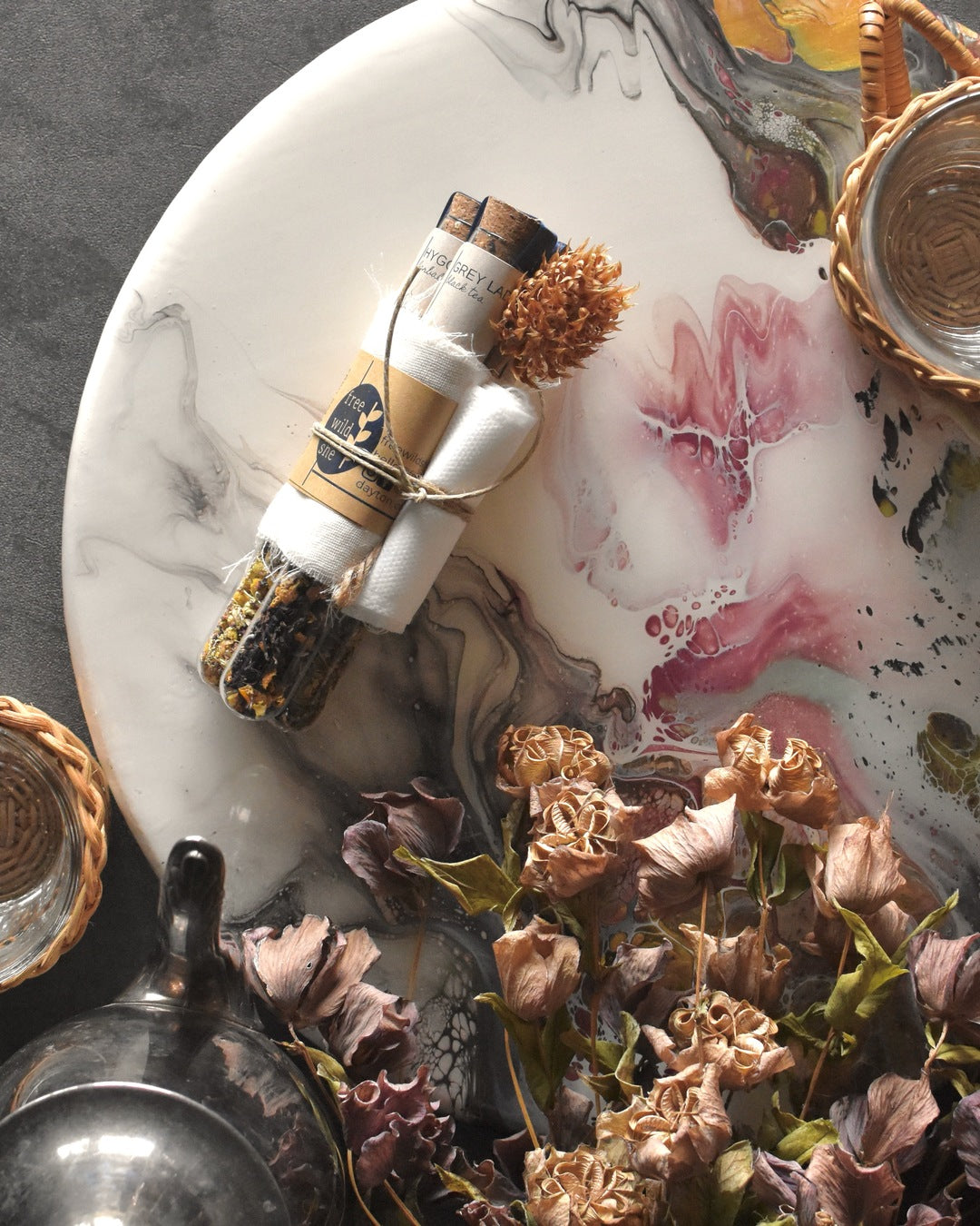Three FreeWildShe tea vials are bundled together with a wrap of muslin, kraft paper, & twine. A sprig of dried clover is tucked into the bundle. The Mother's Mix tea gift pack sits on a platter surrounded by glass & wicker tea mugs, a metal teapot, and dried flowers.