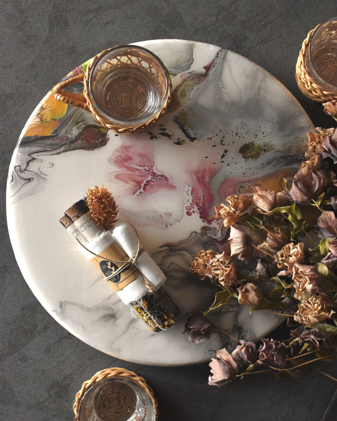 Three FreeWildShe tea vials are bundled together with a wrap of muslin, kraft paper, & twine. A sprig of dried clover is tucked into the bundle. The Mother's Mix tea gift pack sits on a platter surrounded by glass & wicker tea mugs and dried flowers.