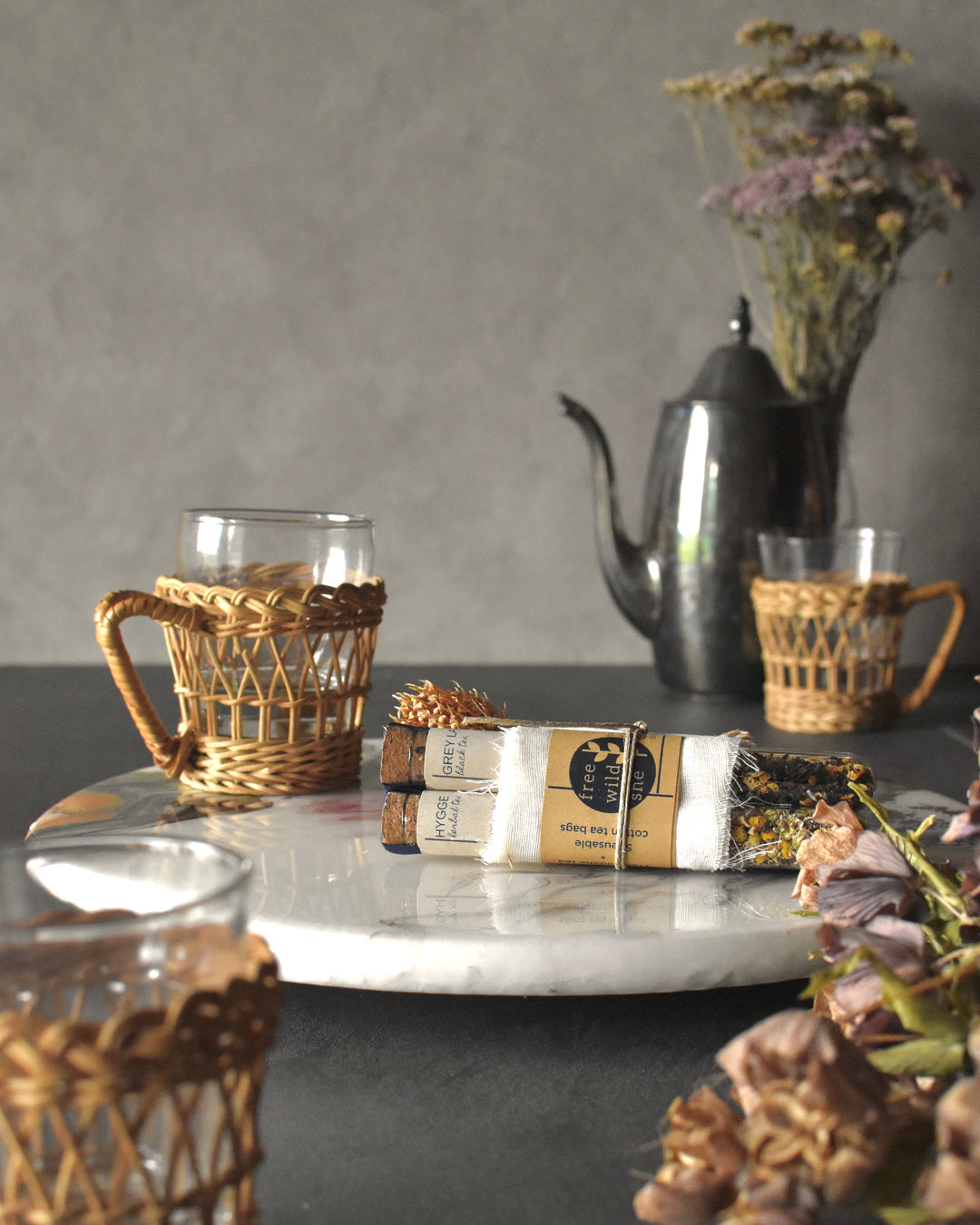 Three FreeWildShe tea vials are bundled together with a wrap of muslin, kraft paper, & twine. A sprig of dried clover is tucked into the bundle. The Mother's Mix tea gift pack sits on a platter surrounded by glass & wicker tea mugs, a metal teapot, and dried flowers.
