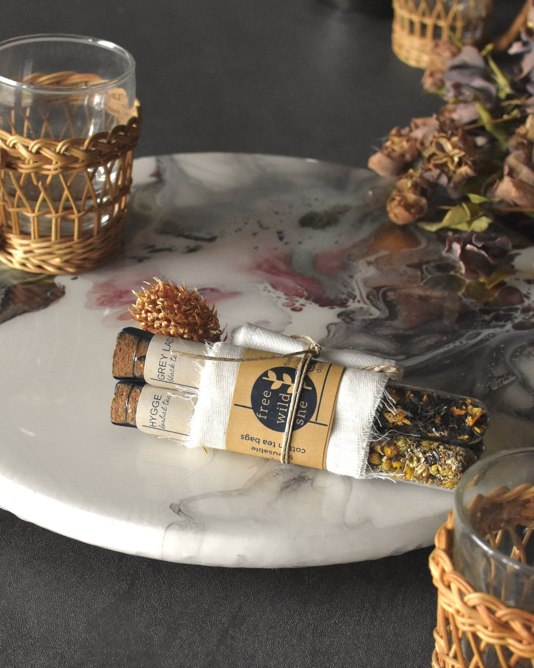 Three FreeWildShe tea vials are bundled together with a wrap of muslin, kraft paper, & twine. A sprig of dried clover is tucked into the bundle. The Mother's Mix tea gift pack sits on a platter surrounded by glass & wicker tea mugs and dried flowers.