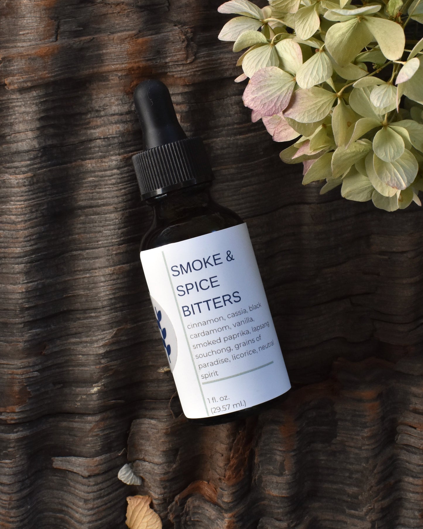 FreeWildShe Smoke & Spice Bitters lay on a textured piece of bark surrounded by dried hydrangea blooms.