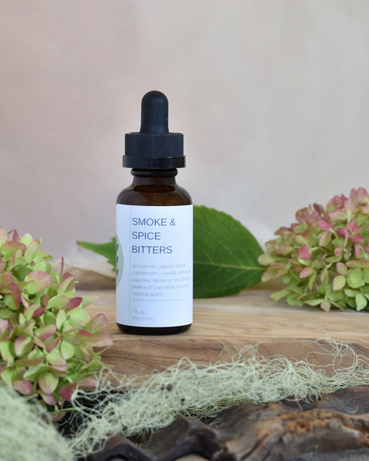 FreeWildShe Smoke & Spice Bitters sit on a wooden tray. Lichen and hydrangea blooms surround the image.