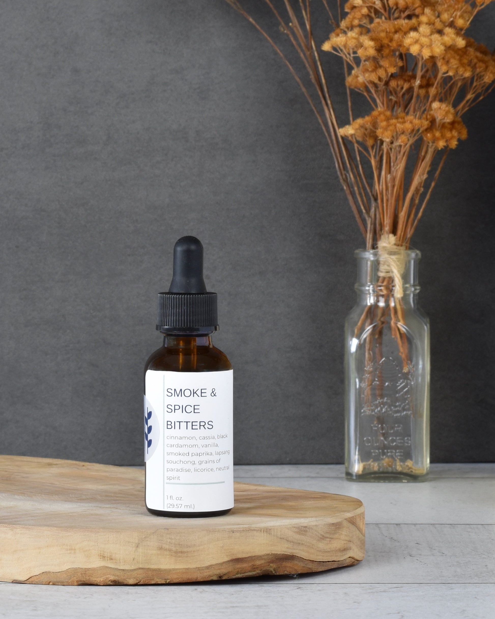 FreeWildShe Smoke & Spice Bitters sit on a wooden platter, on a white wooden table. A bouquet of dried flowers is in the background.