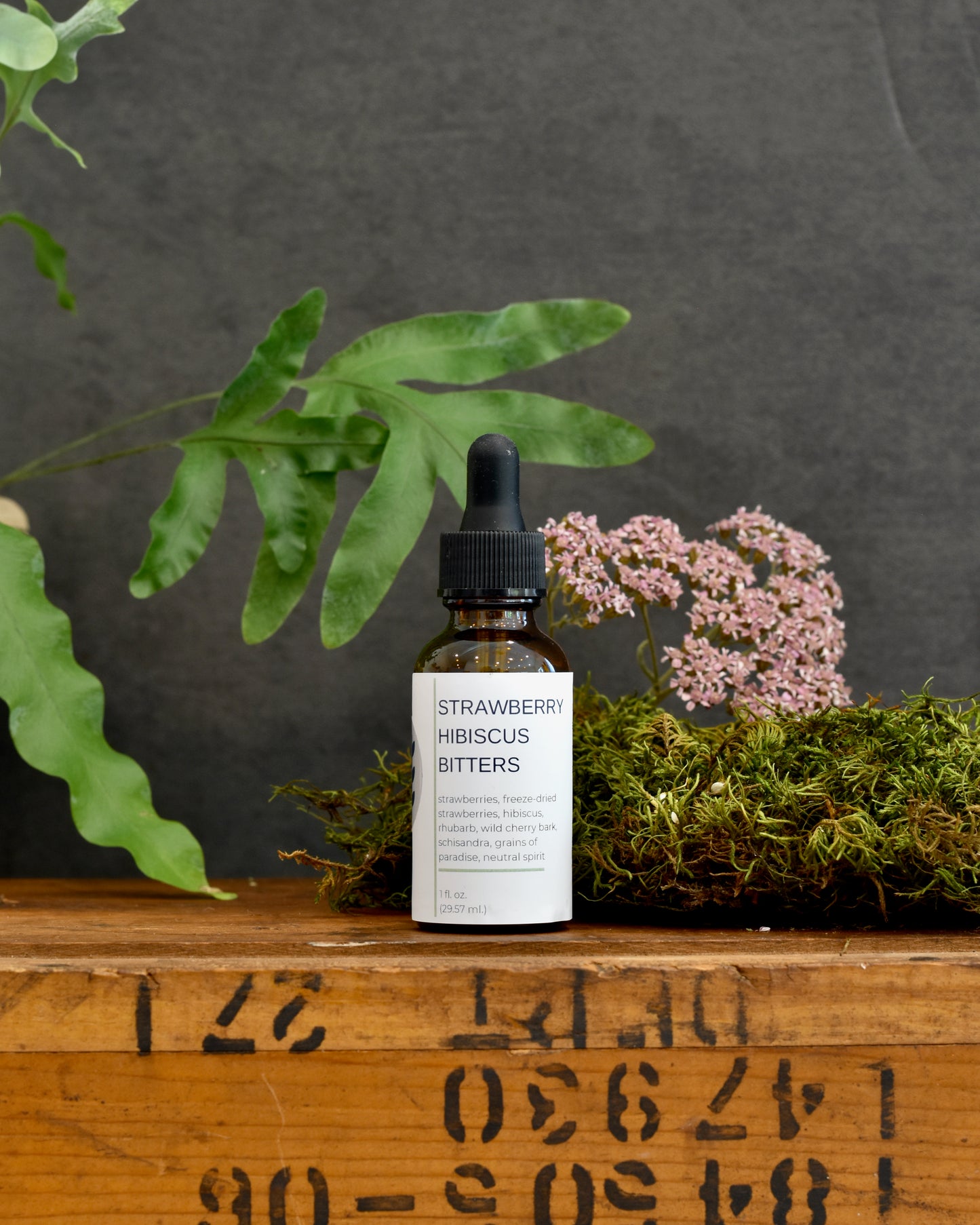 FreeWildShe Strawberry Hibiscus Bitters sit on a wooden box. A plant and dried flower sit in the background.