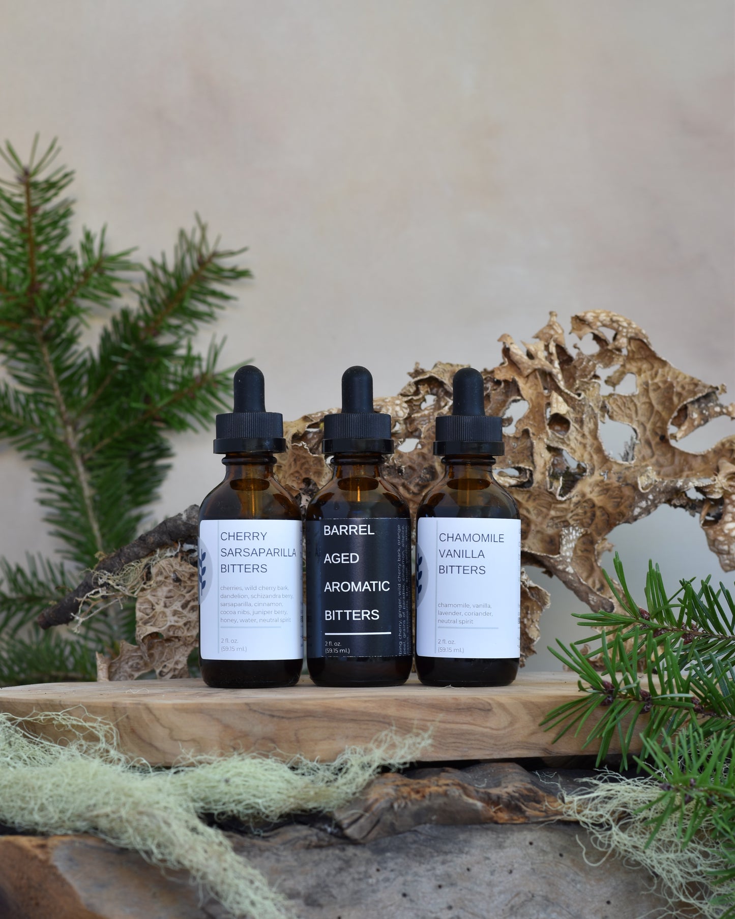 The whiskey lover's bitters bundle sits on a wooden tray surrounded by lichen & douglas fir. From left to right, his trio includes: Cherry Sarsaparilla Bitters, Barrel Aged Aromatic Bitters, and Chamomile Vanilla Bitters.