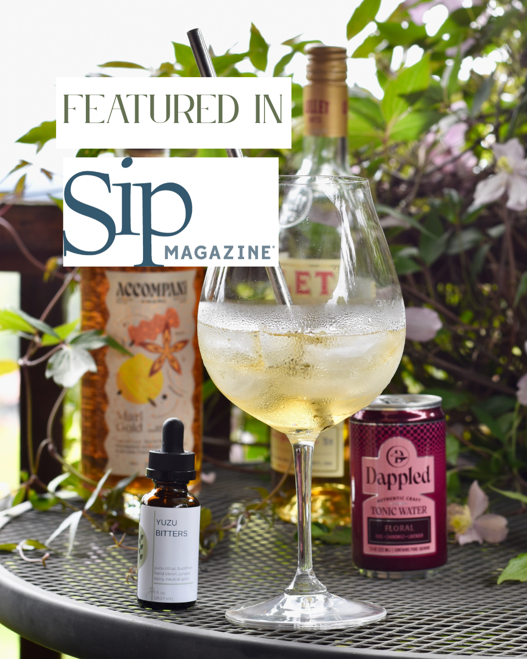 A Yuzu in the Sun cocktail and FreeWildShe Yuzu Bitters sit on a table outside. They are surrounded by ingredients and a green & pink flowering vine. There is a text overlay reading "Featured In Sip Magazine".