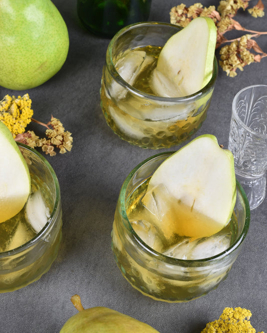 Three Au Pear cocktails sit on a table surrounded by dried botanicals, an etched glass jigger, and fresh green pears. Each drink has a slice of fresh pear to garnish.