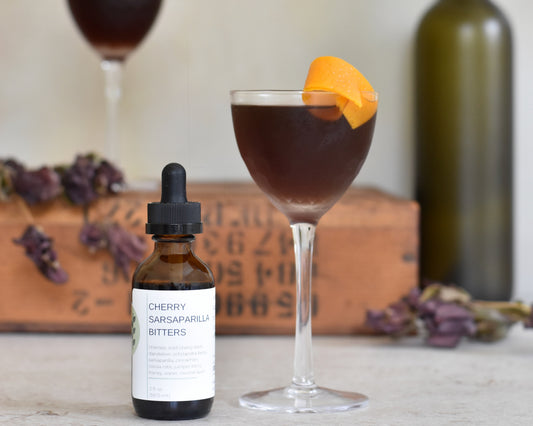 A bottle of FreeWildShe Cherry Sarsaparilla Bitters sits next to a Black Thicket cocktail. Another cocktail sits on a wooden box in the background. There are deep purple dried flowers and a green bottle in the background.