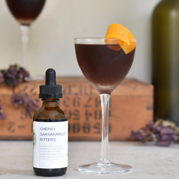 A bottle of FreeWildShe Cherry Sarsaparilla Bitters sits next to a Black Thicket cocktail. Another cocktail sits on a wooden box in the background. There are deep purple dried flowers and a green bottle in the background.