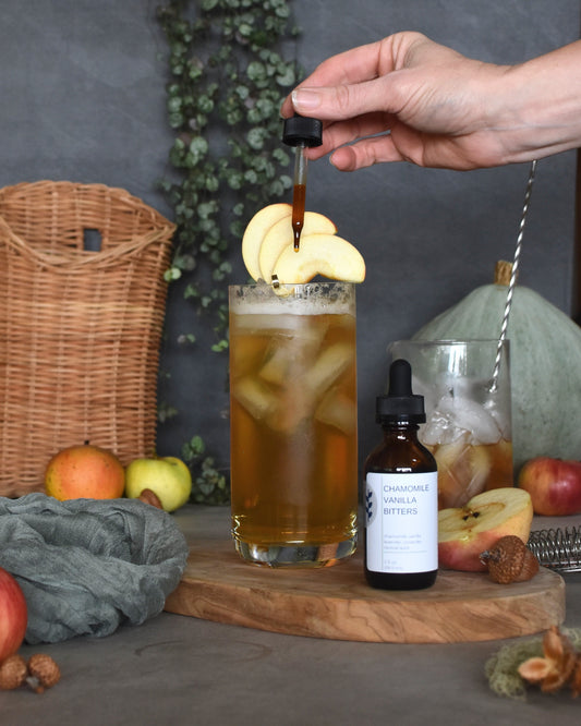 A Honey Pie fall bourbon cocktail sits on a wooden platter. Chamomile Vanilla Bitters sit next to the drink and a hand is reaching in from the side dropping in the bitters. Apples, a giant pale green squash, a basket, and houseplant surround the scene.