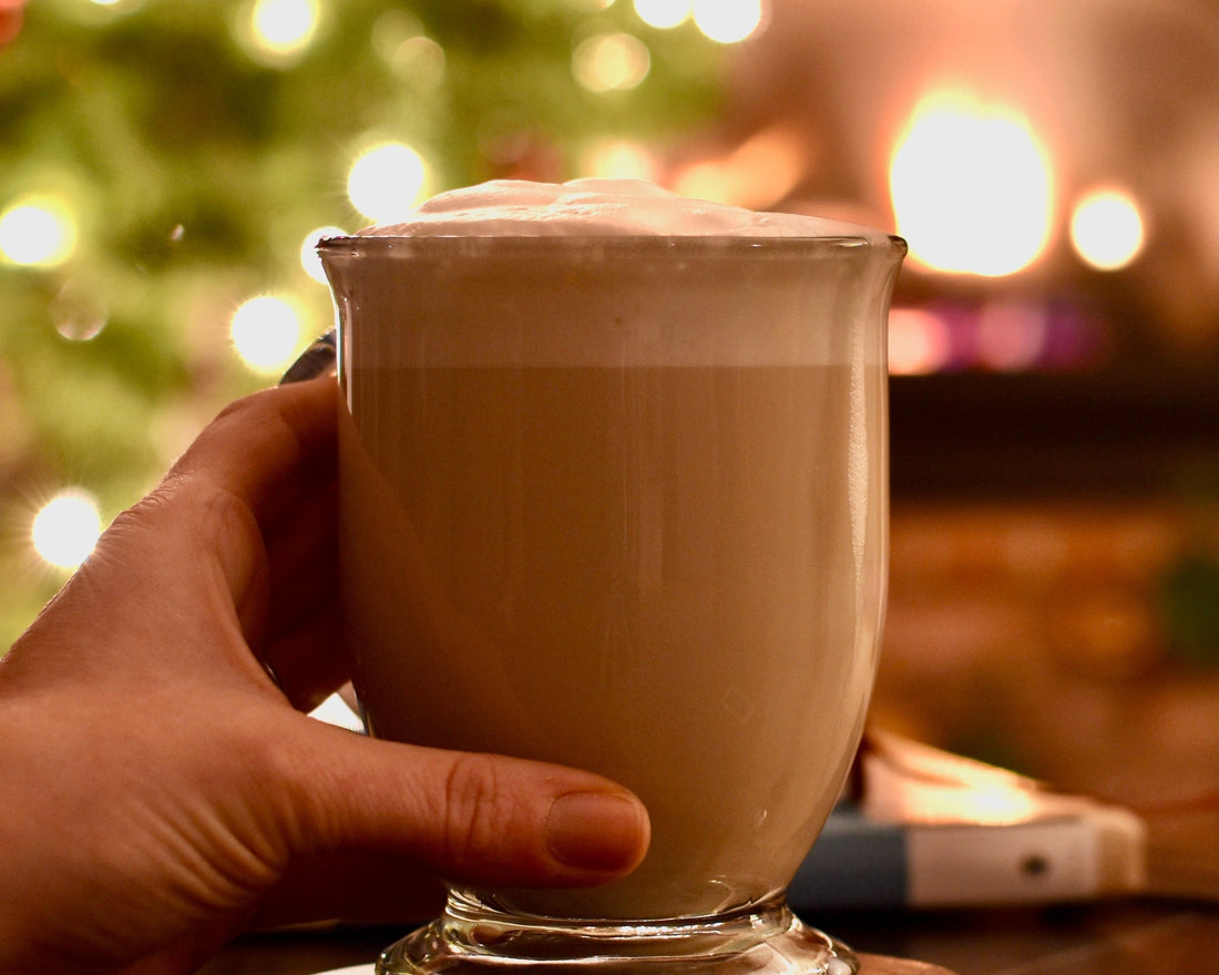 A glass mug of Long Night's Moon Milk sits on a table. A hand gently wraps around. The blur of a Christmas tree & fireplace are in the background.