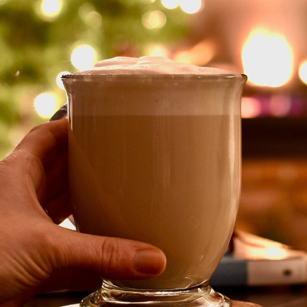 A glass mug of Long Night's Moon Milk sits on a table. A hand gently wraps around. The blur of a Christmas tree & fireplace are in the background.