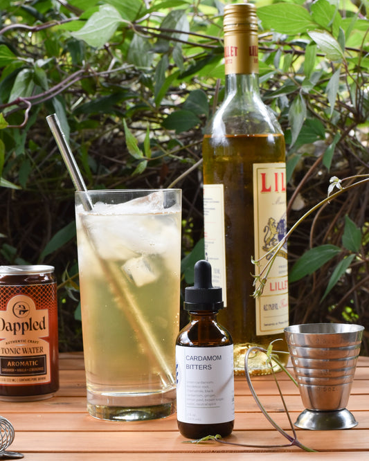 FreeWildShe Cardamom Bitters sit in front of a Porch Swing summer whiskey cocktail on a wooden table outside. Ingredients and bar tools are scattered around. The backdrop is a vibrant green vine.