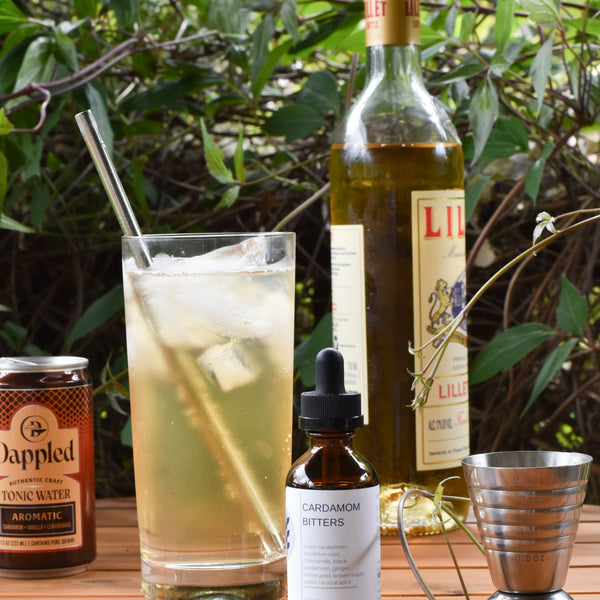 FreeWildShe Cardamom Bitters sit in front of a Porch Swing summer whiskey cocktail on a wooden table outside. Ingredients and bar tools are scattered around. The backdrop is a vibrant green vine.