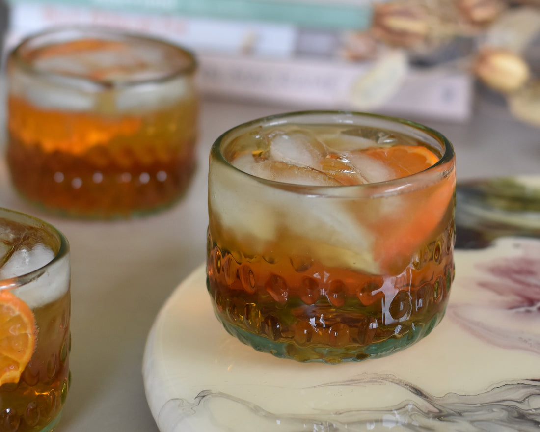 The Wayward Orchard cocktail by FreeWildShe sits on a pastel watercolor tray. The drink is a warm cola brown color with a vibrant orange slice and the glass is dimpled. Two more cocktails, a stack of books, and dried flowers sit in the background.