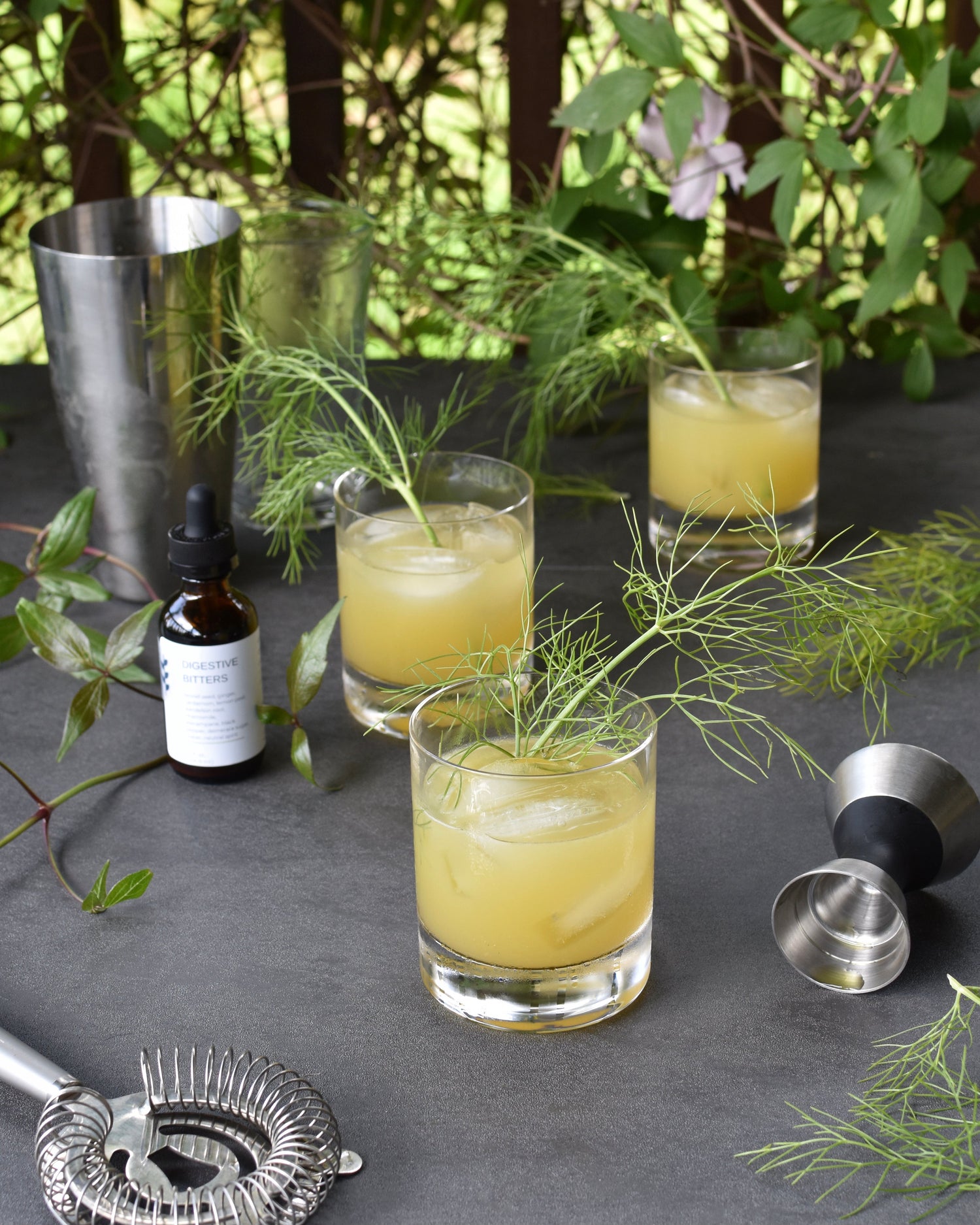 Three Verdant Horizon pineapple gin cocktails sit on a table outside. They are surrounded by FreeWildShe Digestive Bitters, bar tools, and vibrant green leaves.