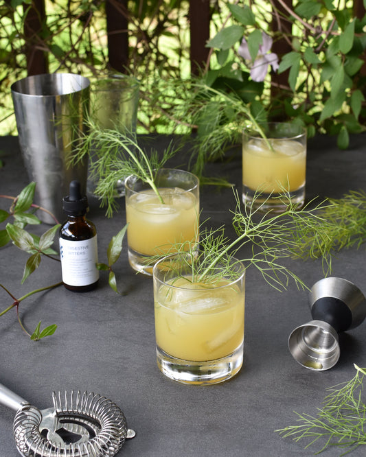 Three Verdant Horizon pineapple gin cocktails sit on a table outside. They are surrounded by FreeWildShe Digestive Bitters, bar tools, and vibrant green leaves.