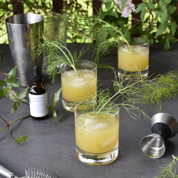 Three Verdant Horizon pineapple gin cocktails sit on a table outside. They are surrounded by FreeWildShe Digestive Bitters, bar tools, and vibrant green leaves.