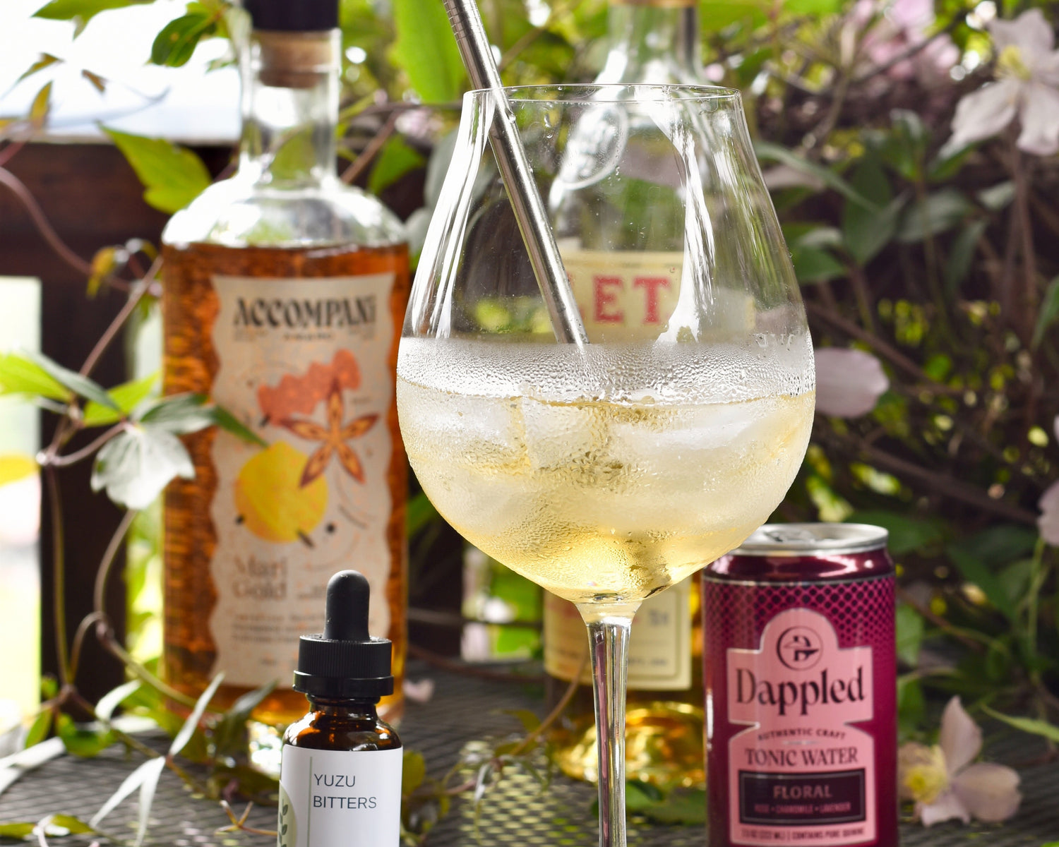 A Yuzu in the Sun cocktail sits on a table outside. It is surrounded by ingredients and a green vine.