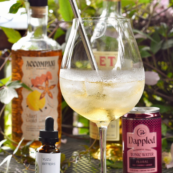 A Yuzu in the Sun cocktail sits on a table outside. It is surrounded by ingredients and a green vine.