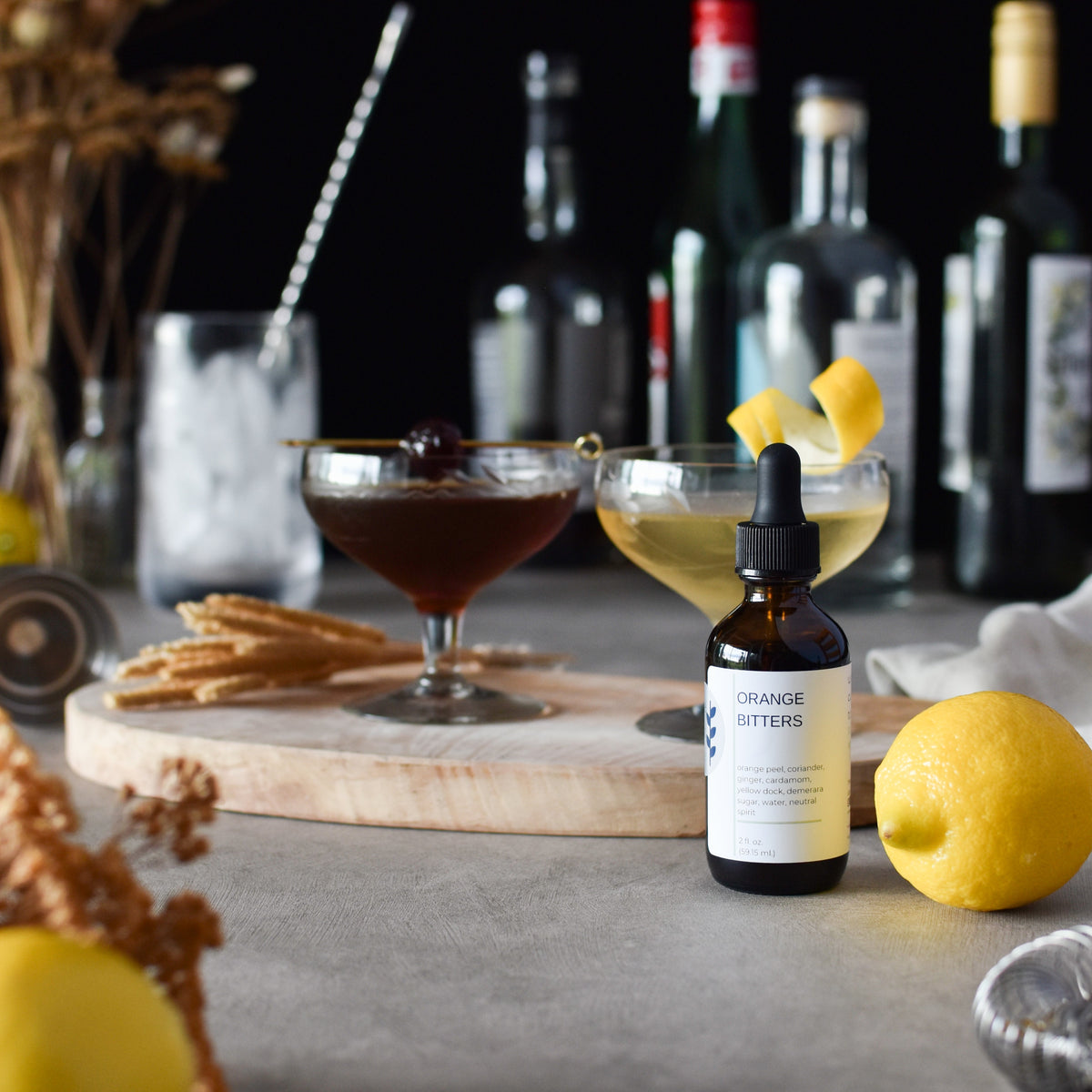 An amber glass dropper bottle labeled 'ORANGE BITTERS' stands on a gray tabletop beside two cocktails, a lemon, and blurred bottles in the background.