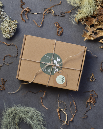 A brown box with an olive green sticker that reads Free Wild She on the left with a leaf image on the right. The box is wrapped with twine and has a gift tag. Lichen and brown crinkle stuffing are scattered around the box.