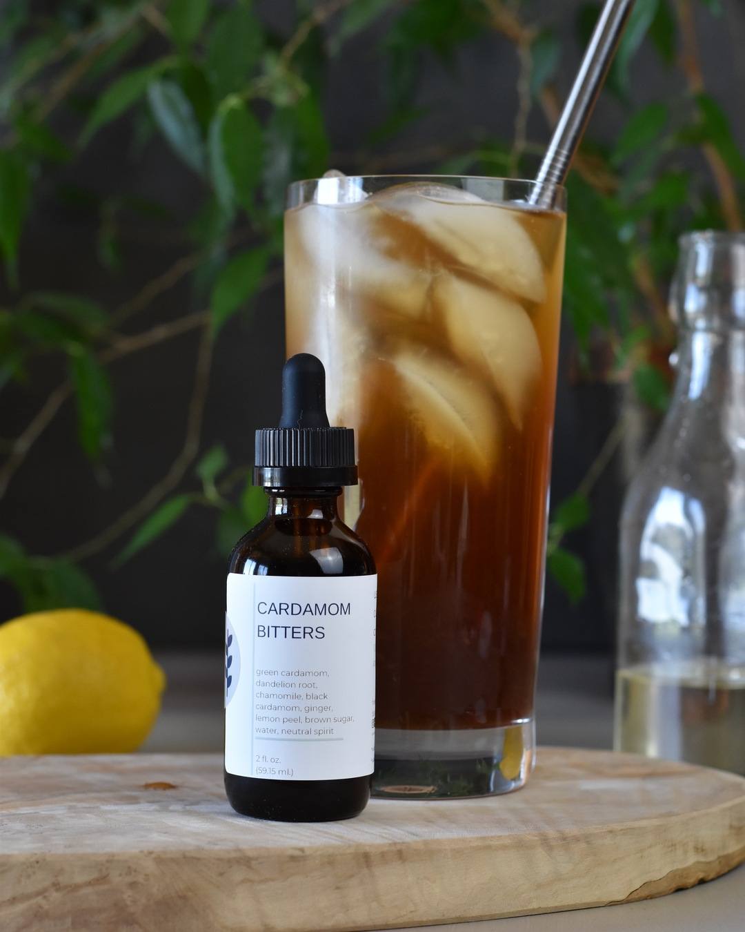 FreeWildShe Cardmom Bitters sit in front of a Cardamom Coffee Spritzer on a wooden platter. A lemon, simple syrup, and houseplant sit in the background.