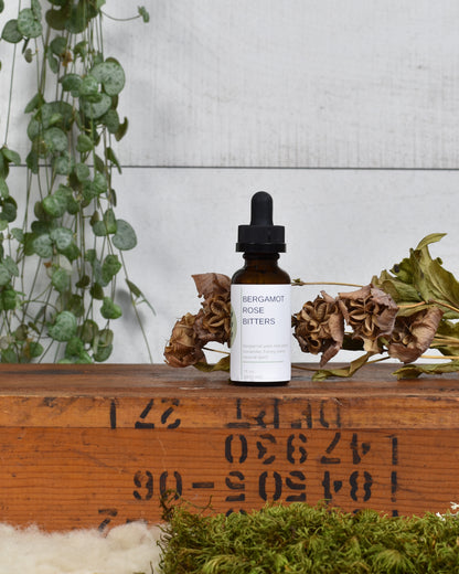 FreeWildShe Bergamot Rose Bitters sit on a wooden box. Dried flowers & moss sit in front of the box and a houseplant hangs behind.