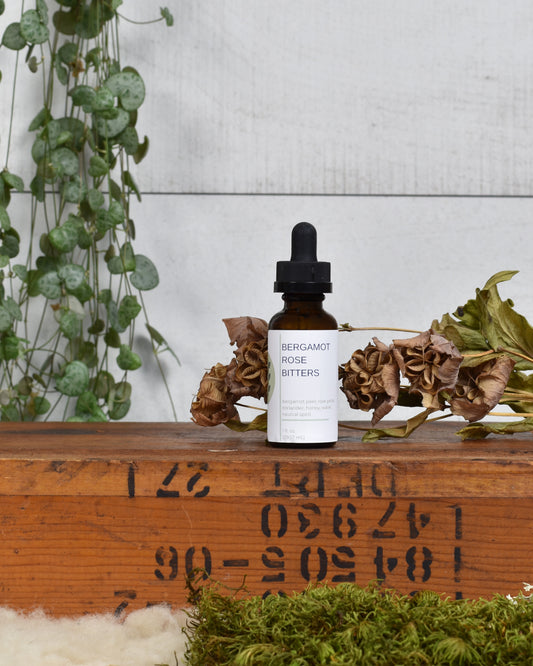 FreeWildShe Bergamot Rose Bitters sit on a wooden box. Dried flowers & moss sit in front of the box and a houseplant hangs behind.