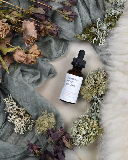 FreeWildShe Bergamot Rose Bitters lay on green fabric surrounded by dried flowers & lichen.