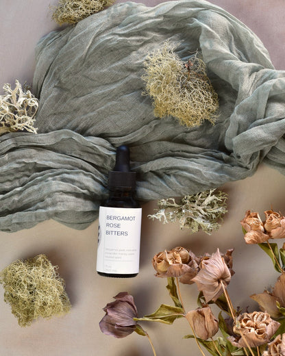 FreeWildShe Bergamot Rose Bitters lay on green fabric surrounded by dried flowers & lichen.