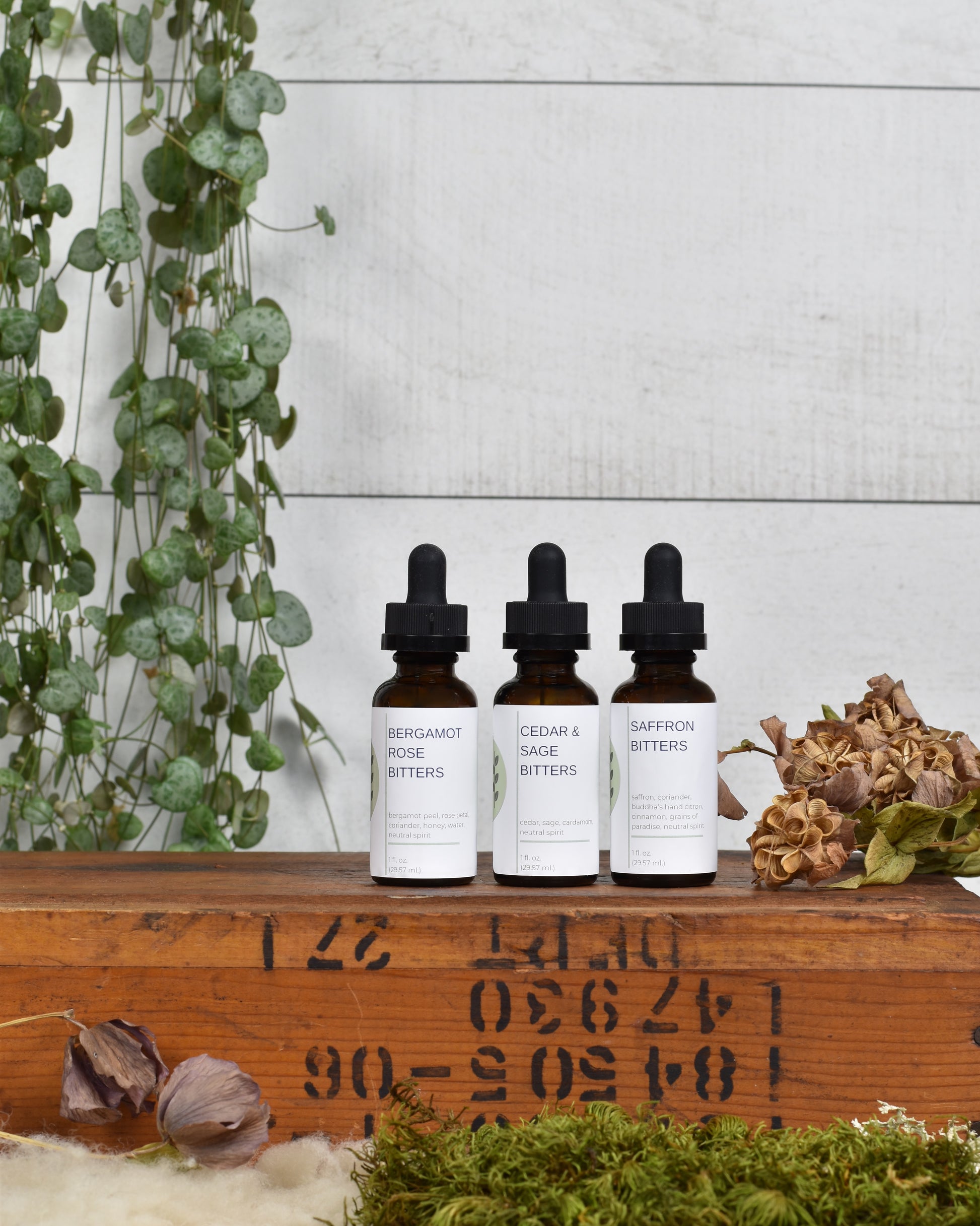 FreeWildShe Limited Bitters Collection featuring Bergamot Rose, Cedar & Sage, and Saffron sit on a wooden box. Dried flowers & moss sit in front of the box and a houseplant hangs behind.