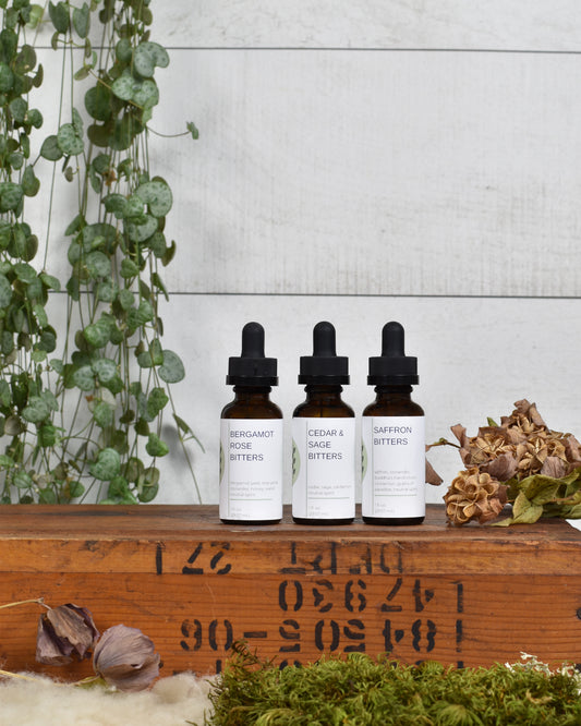 FreeWildShe Limited Bitters Collection featuring Bergamot Rose, Cedar & Sage, and Saffron sit on a wooden box. Dried flowers & moss sit in front of the box and a houseplant hangs behind.