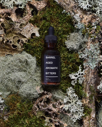 FreeWildShe x Bull Run Distillery Barrel-Aged Aromatic Bitters sit on moss & lichen.