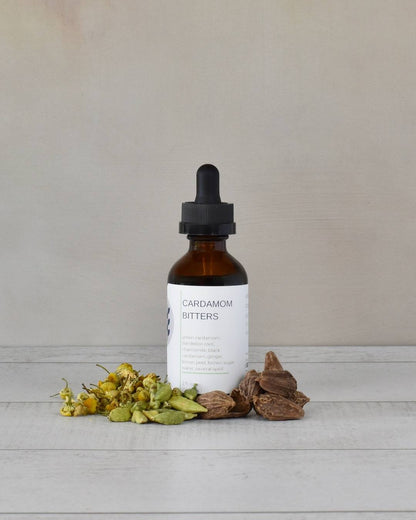 FreeWildShe Cardamom Bitters sit on a white countertop surrounded by ingredients.