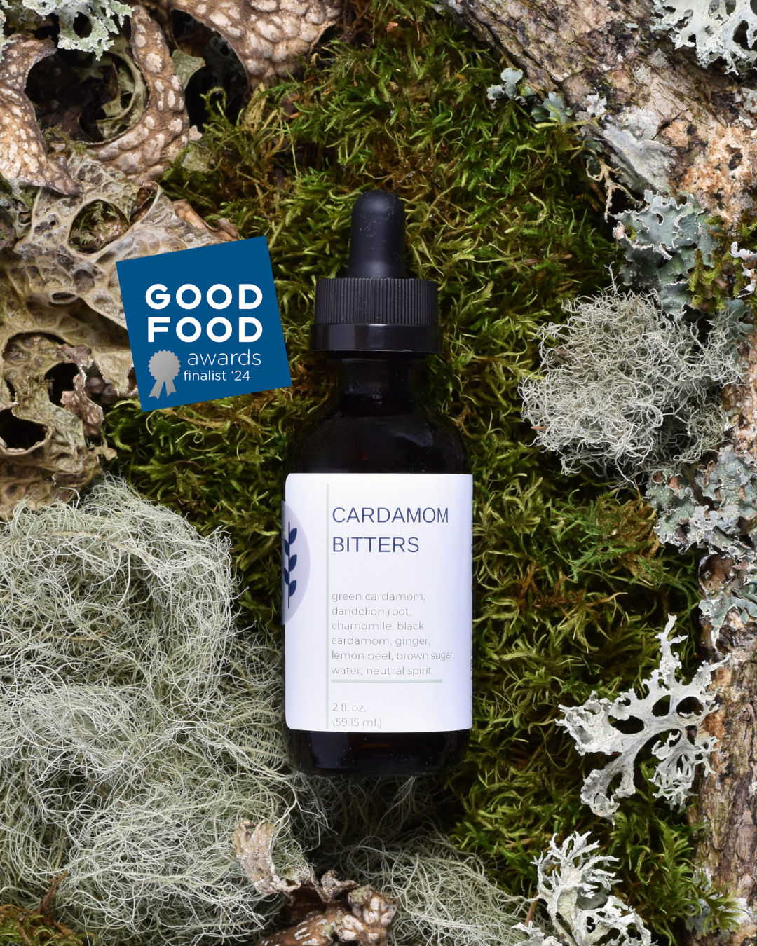 FreeWildShe Cardamom Bitters lay on a bed of moss & lichen. A navy blue, square badge that reads, 'Good Food Awards Finalist '24' sits on the top left side.