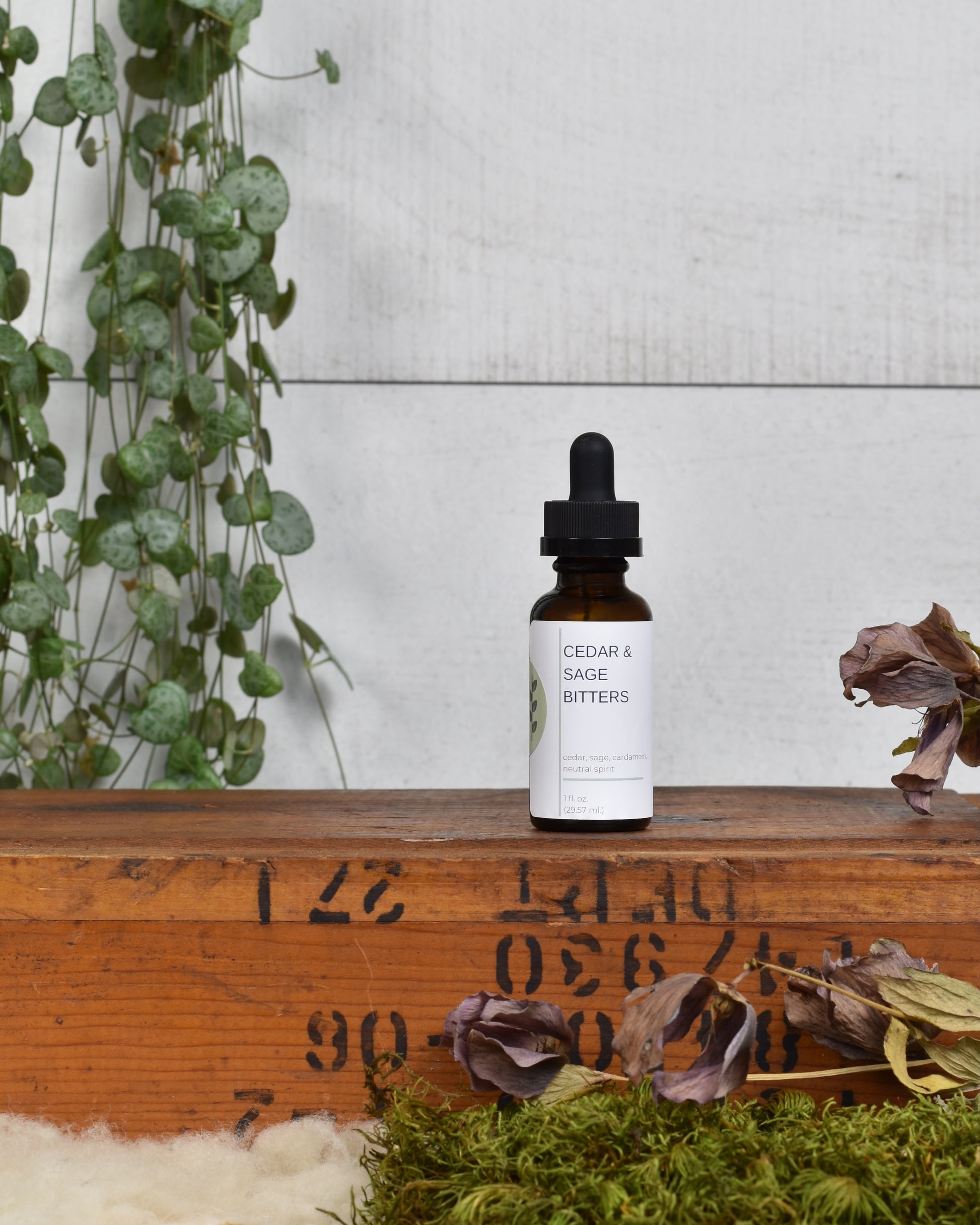 FreeWildShe Cedar & Sage Bitters sit on a wooden box. Dried flowers & moss sit in front of the box and a houseplant hangs behind.