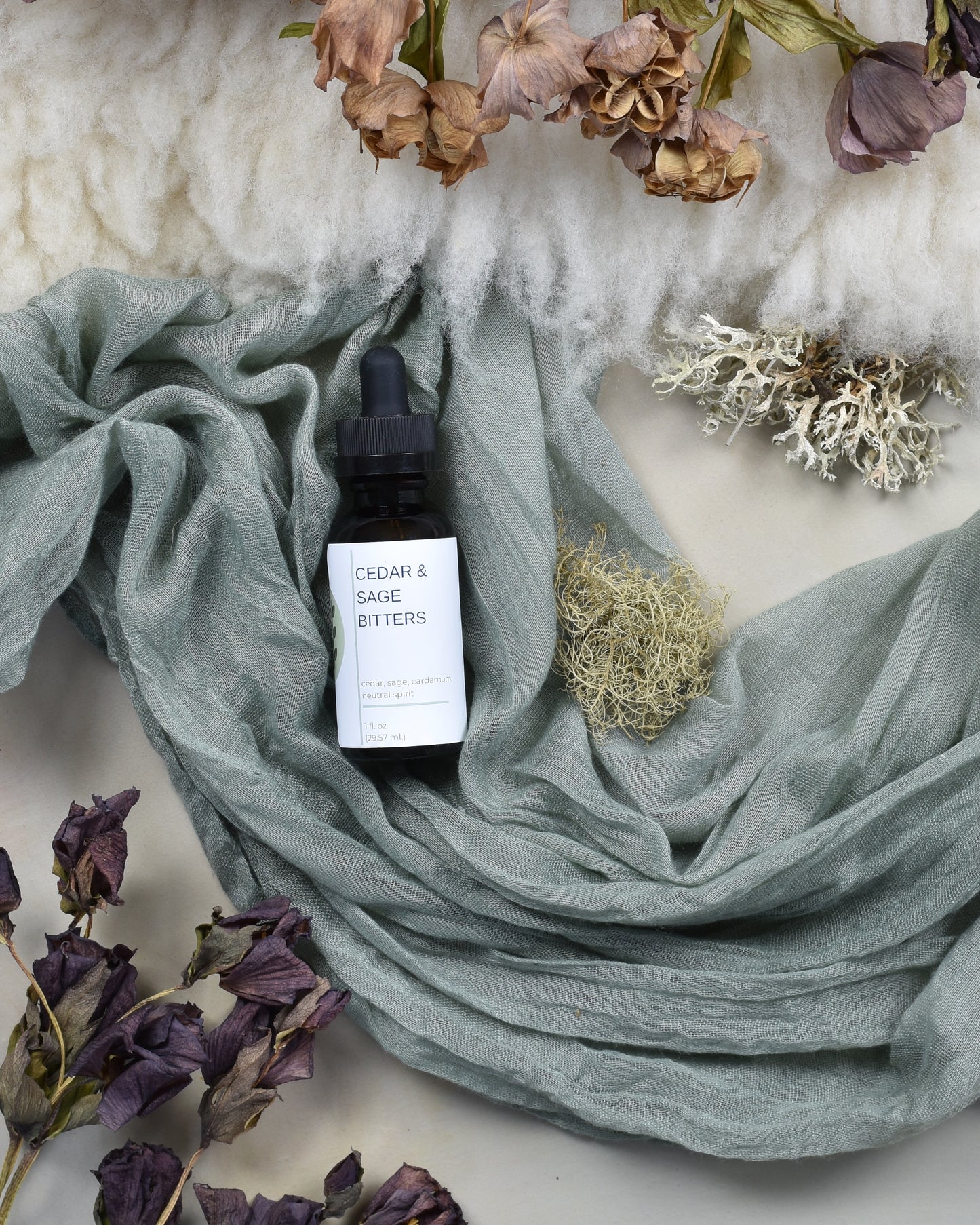FreeWildShe Cedar & Sage Bitters lay on green fabric surrounded by dried flowers & lichen.
